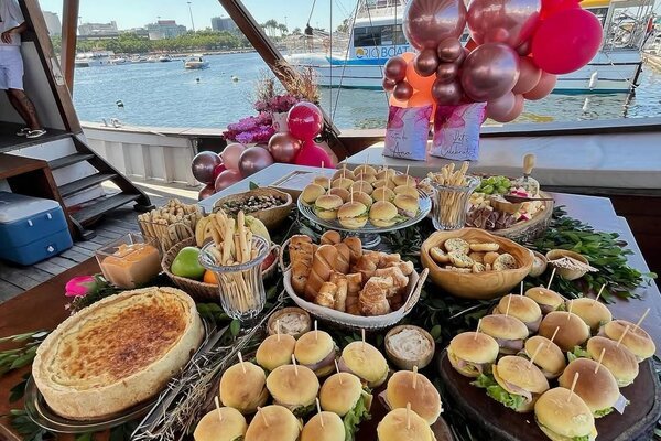 Mesa de buffet com mini hambúrgueres, quiche e salgados decorada com balões rosa em festa de aniversário na lancha